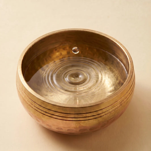 Multirow row 3 hero: cymatics mechanism — bronze singing bowl with single suspended droplet and concentric ripple pattern